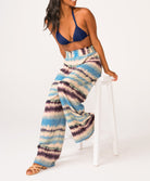 A woman wearing a navy triangle bikini top and blue tie dye pants sitting on a white stool.