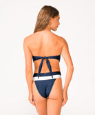 A woman wearing a navy and white color block bandeau bikini facing towards a white wall.