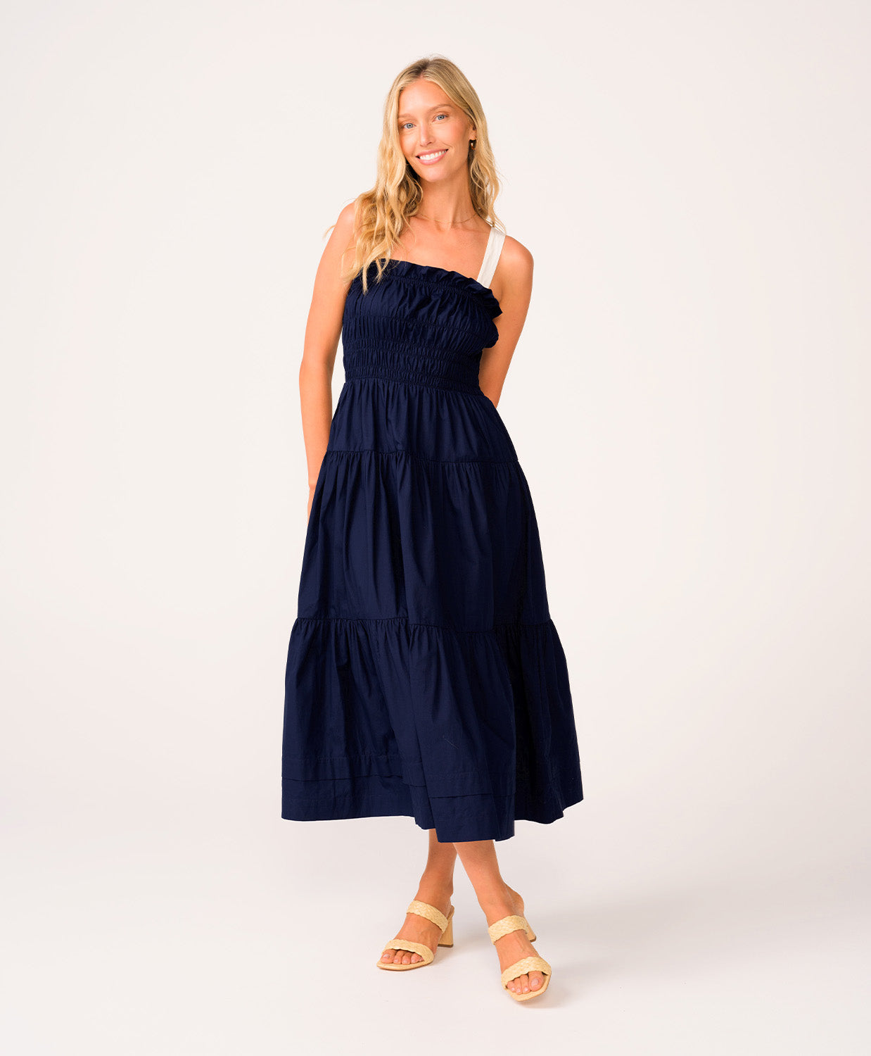 A woman wearing a navy dress with white straps standing against a white wall.