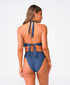 A woman wearing a navy halter style bikini top and matching bottoms facing towards a white wall.