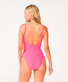 A woman wearing a pink one piece bathing suit with an open back and adjustable straps facing away in front of a white background.