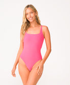 A woman wearing a pink one piece bathing suit standing against a white wall.