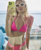 A woman wearing a pink triangle top bikini sitting on a blue chair and holding a green iphone.
