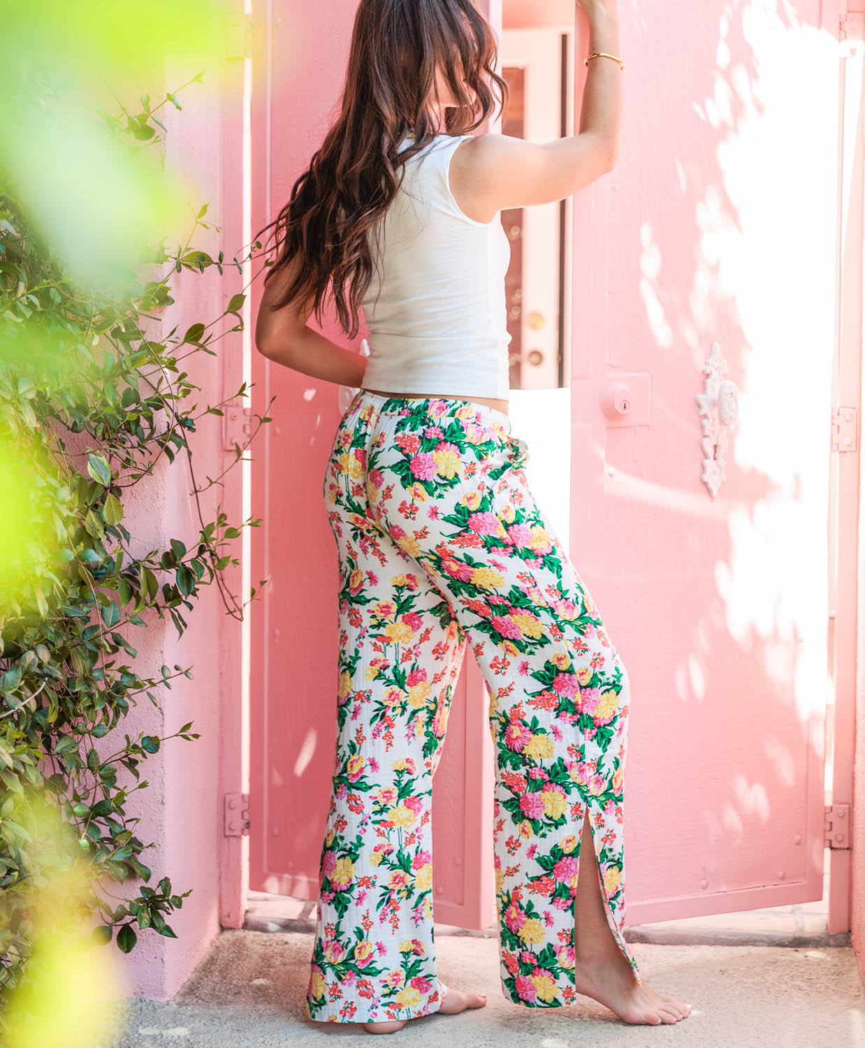 Woman wearing a white top and colorful floral pants standing against a pink wall with greenery.