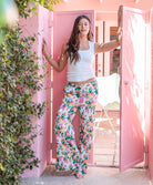 Woman in a white top and floral pants standing in front of a pink door.