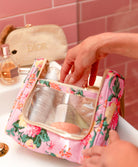 Hand reaching into a floral makeup bag with skincare products on a pink tiled wall background