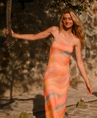 A woman wearing a strapless, shimmering orange maxi dress with wavy pink, peach, and gray-blue horizontal stripes, standing outdoors against a textured stone wall.