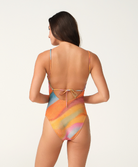 A woman shown from the back wearing a one-piece swimsuit with a colorful abstract print in orange, yellow, pink, blue, and beige tones, photographed against a plain light background.