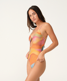 A woman modeling a one-piece swimsuit with a vibrant, abstract wavy pattern in orange, yellow, pink, and light blue on a plain off-white studio background.
