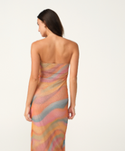 A woman modeling a strapless maxi dress with a wavy, abstract pattern in pastel shades of orange, pink, beige, and light blue, set against a plain light gray studio background.