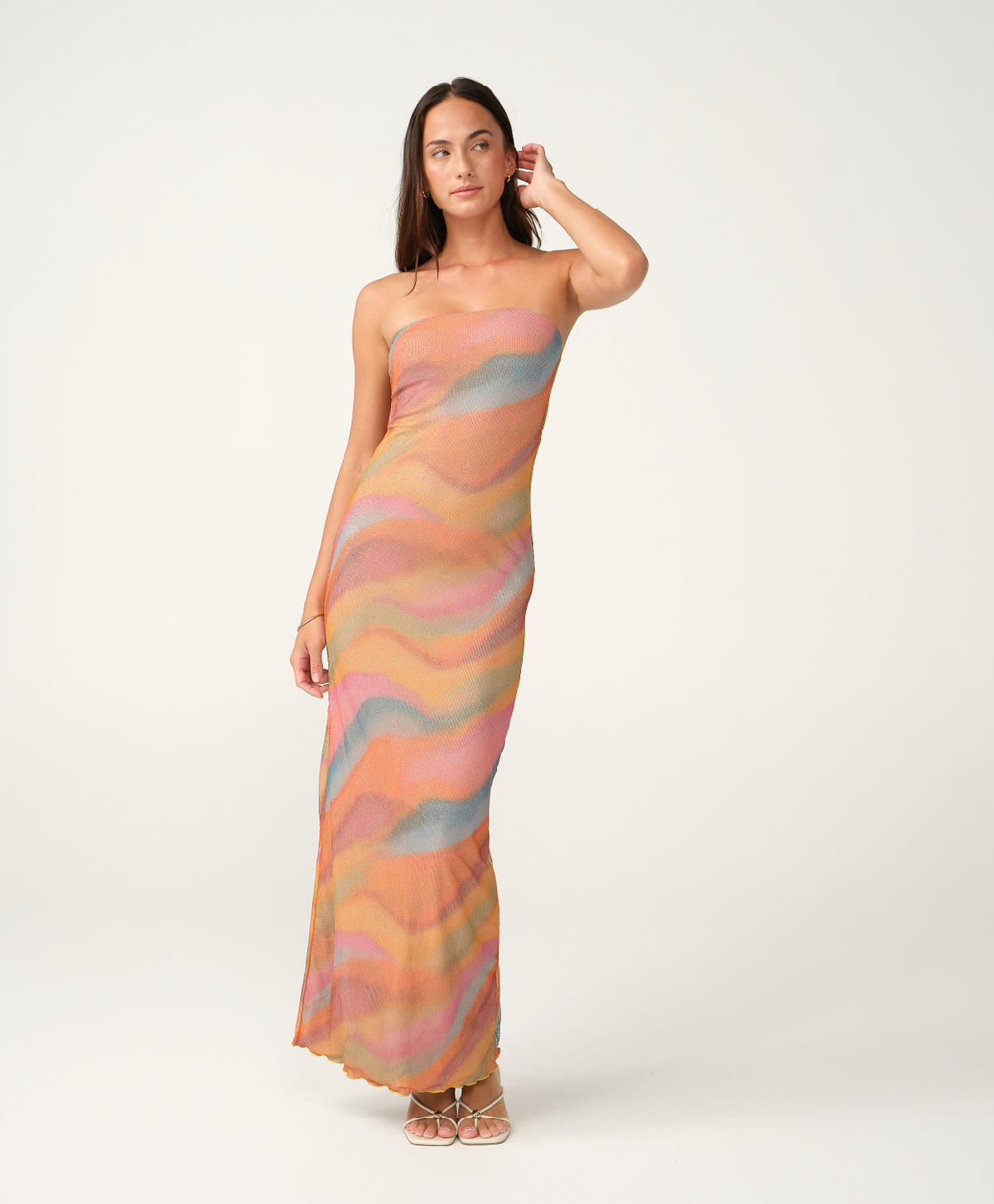 A woman modeling a strapless maxi dress with a wavy, abstract pattern in pastel shades of orange, pink, beige, and light blue, set against a plain light gray studio background.