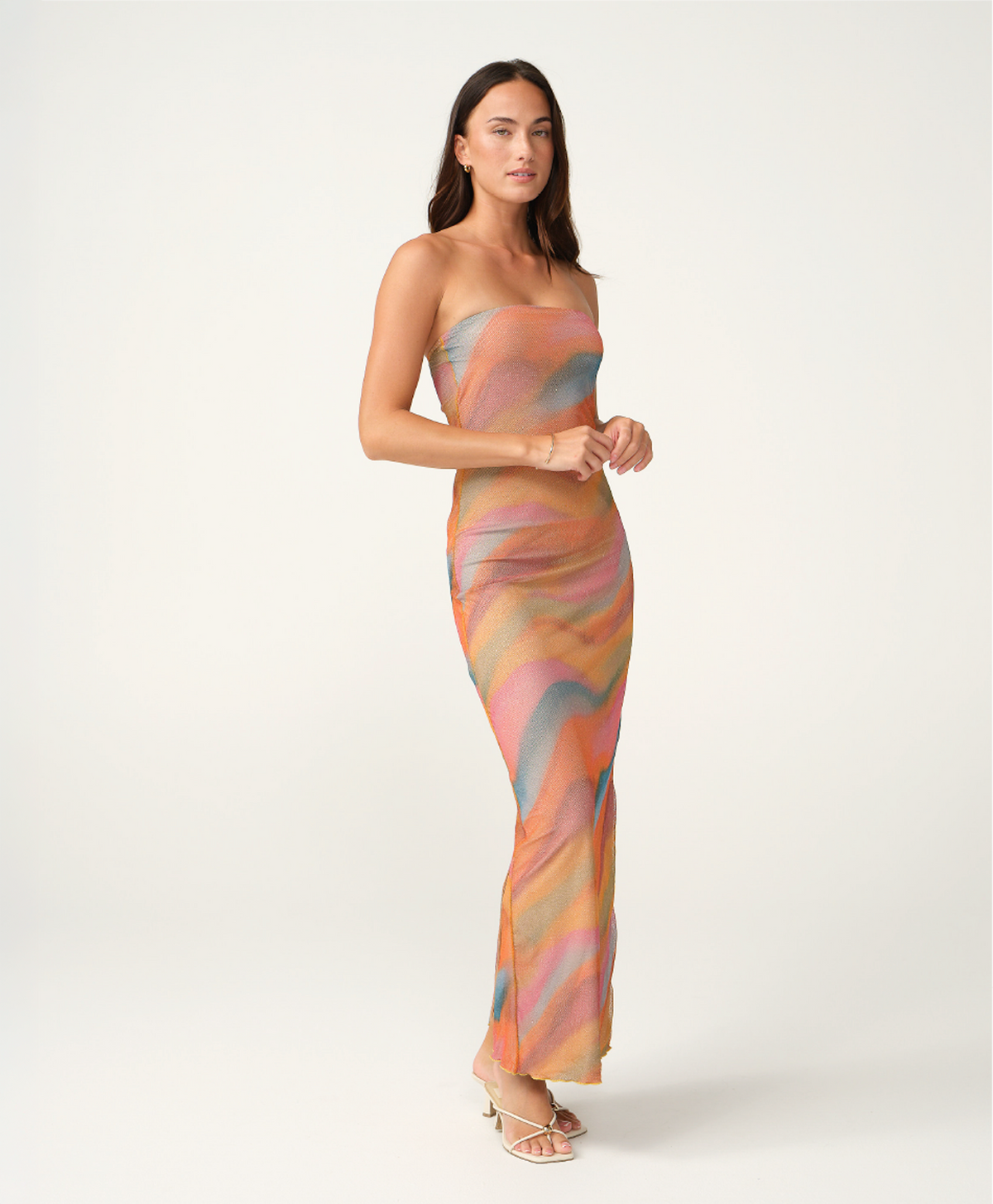 A woman modeling a strapless maxi dress with a wavy, abstract pattern in pastel shades of orange, pink, beige, and light blue, set against a plain light gray studio background.