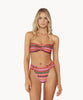 Blonde woman wearing a multi-colored stripe print bandeau bikini with gold details stands in front of white wall.