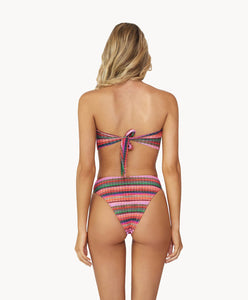Blonde woman wearing a multi-colored stripe print bandeau bikini with gold details facing backwards towards a white wall.