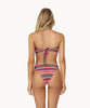 Blonde woman wearing a multi-colored stripe print bandeau bikini with gold details facing backwards towards a white wall.