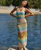A brunette woman wearing a ankle length multi colored dress standing on a rock near the water. 