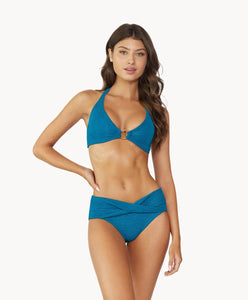 Brunette woman wearing a textured turquoise halter bikini with gold details stands in front of white wall.