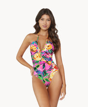 Brunette woman wearing pink tropical print ruched one piece swimsuit stands in front of white wall.