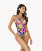 Brunette woman wearing pink tropical print ruched one piece swimsuit stands in front of white wall.