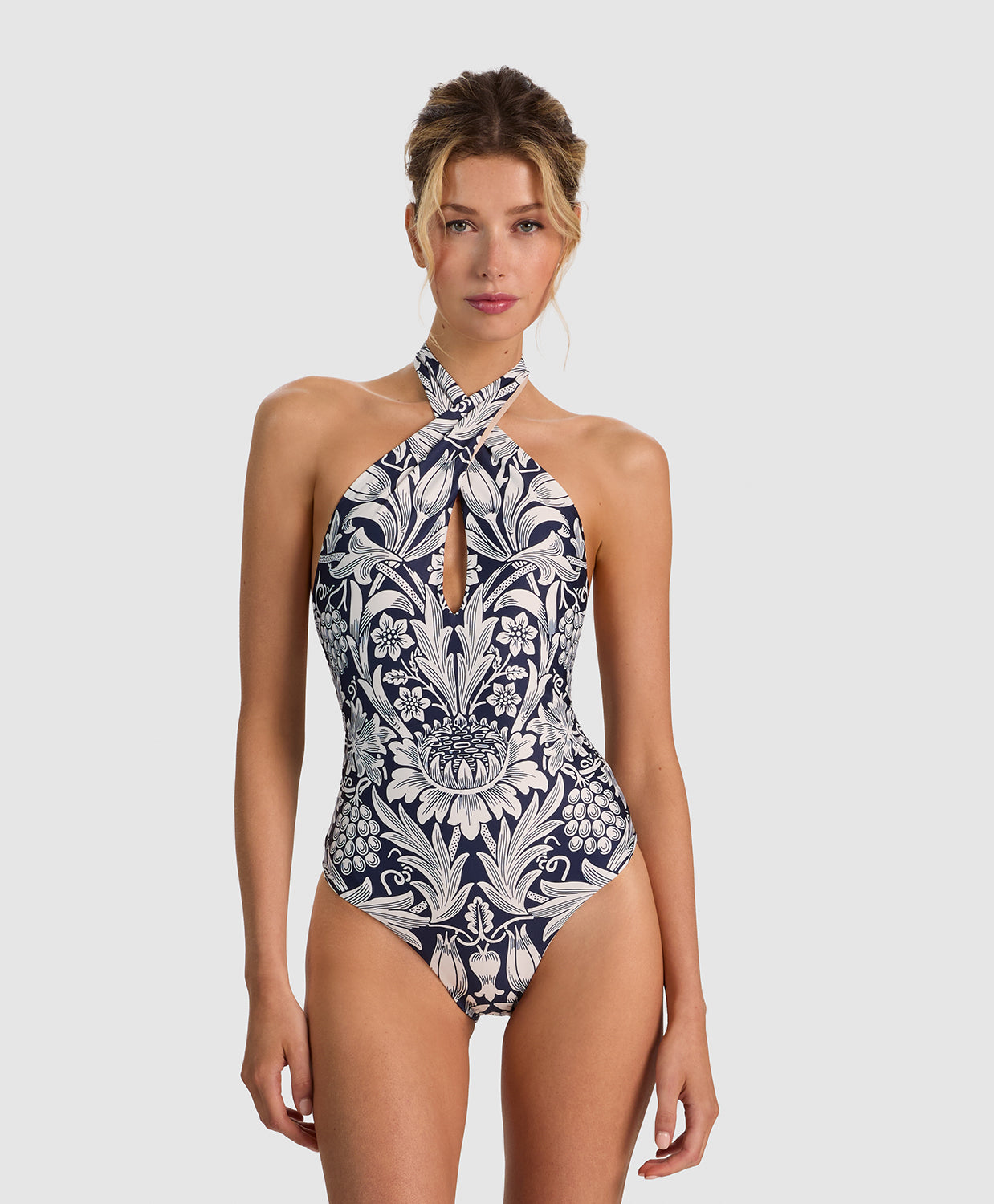 A woman modeling a one-piece halter swimsuit with a navy blue and white botanical pattern, set against a solid light gray background.