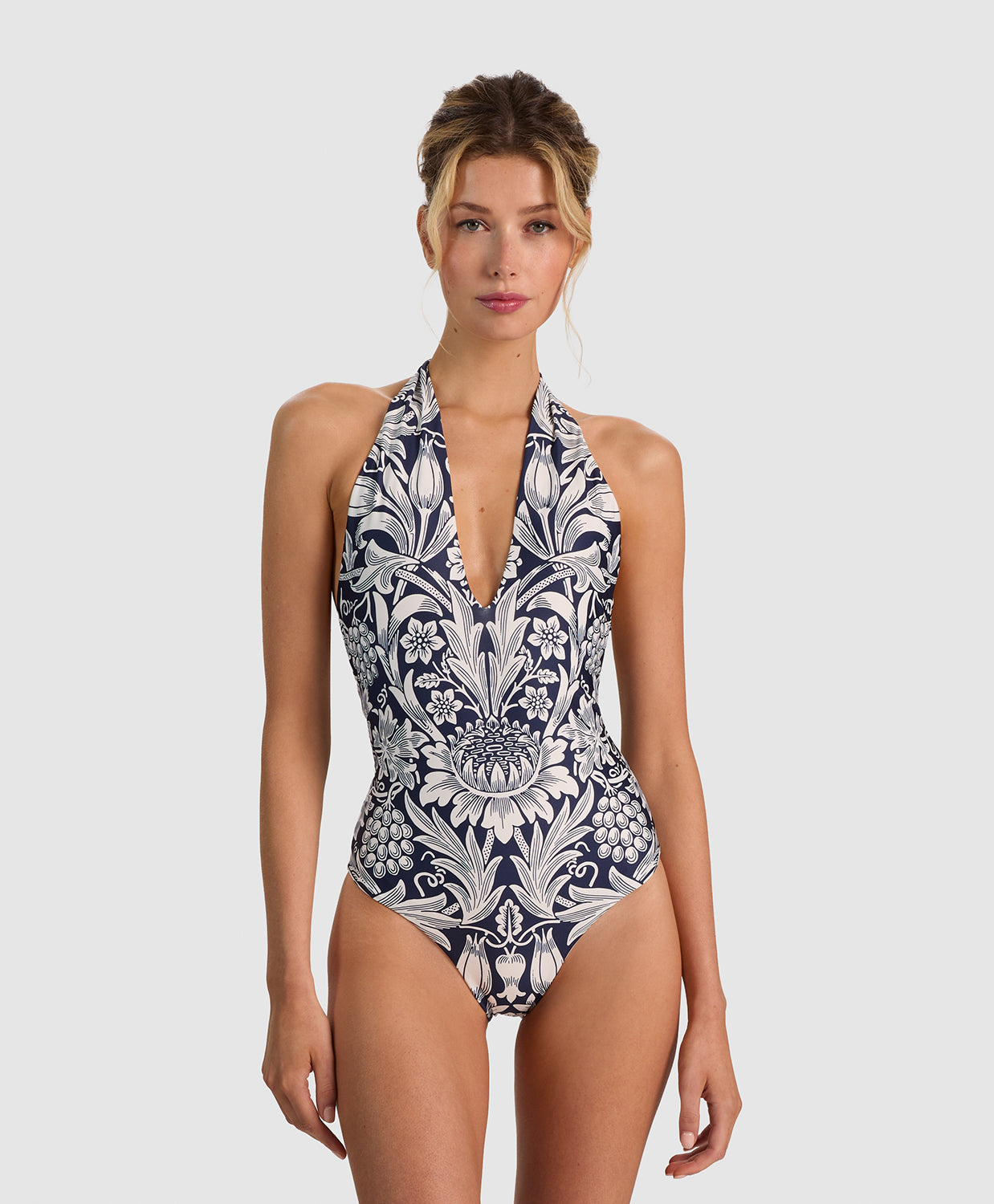A woman modeling a halter-neck one-piece swimsuit with a navy blue and white baroque floral pattern, set against a solid light gray background.