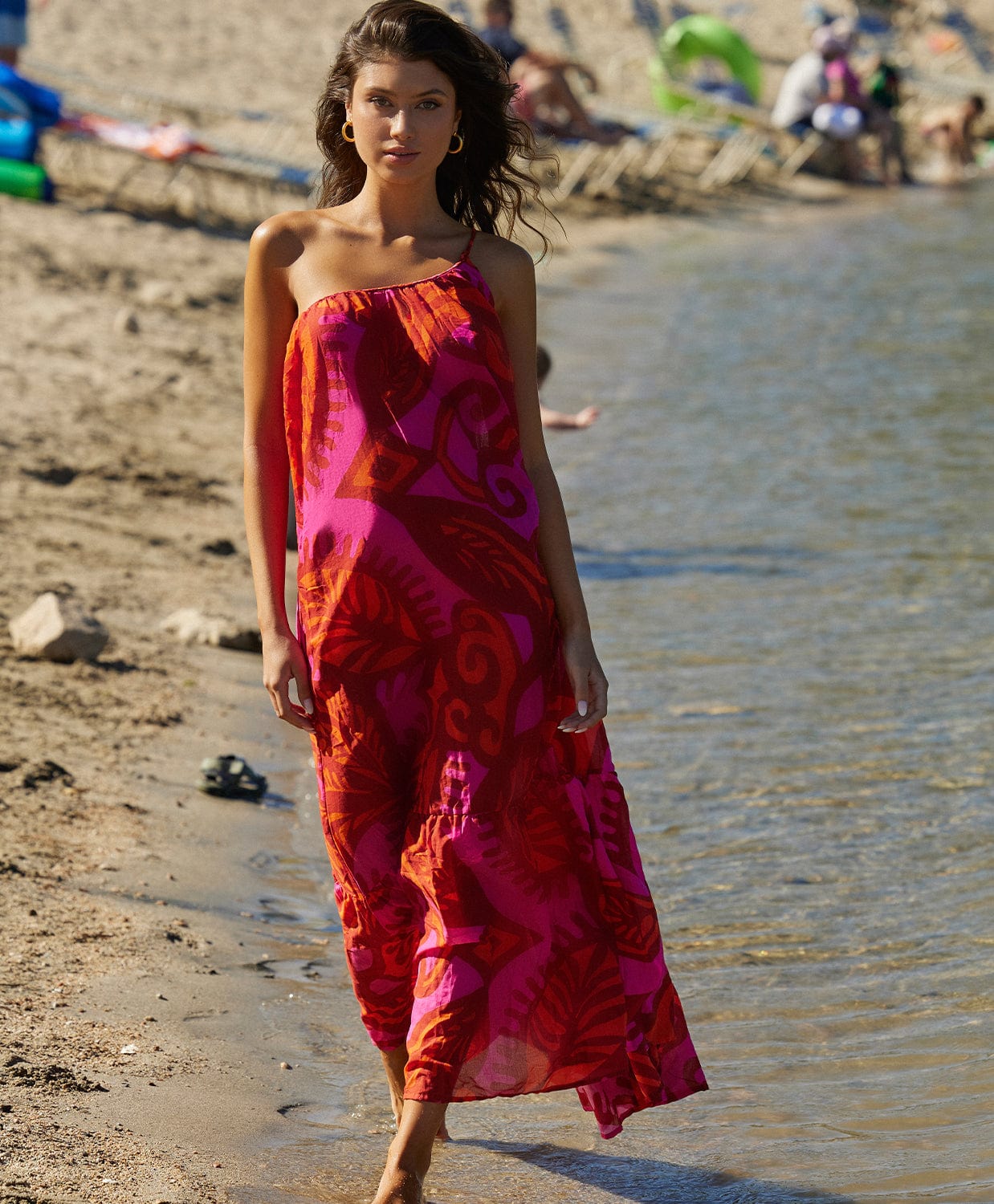 Brunette woman wearing an ankle length dress with one shoulder strap. She is walking on the beach near a lake.