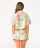 A woman wearing a light blue floral shirt and floral shorts facing towards a white wall.