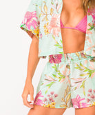 A woman wearing a bright pink halter bikini top and floral shorts standing against a white wall.
