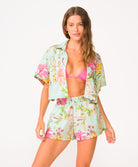 A woman wearing a bright pink halter bikini top and floral shorts standing against a white wall.