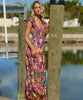 Blonde woman wearing a maxi pink tropical dress with sequins stands on a dock by water.