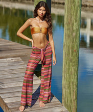 Brunette woman wearing multi-colored stripe print textured pants with a gold bandeau bikini top stands on a pier.