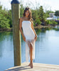 Brunette woman wearing a white strapless fringe hem dress with macramé details stands on a dock by water.