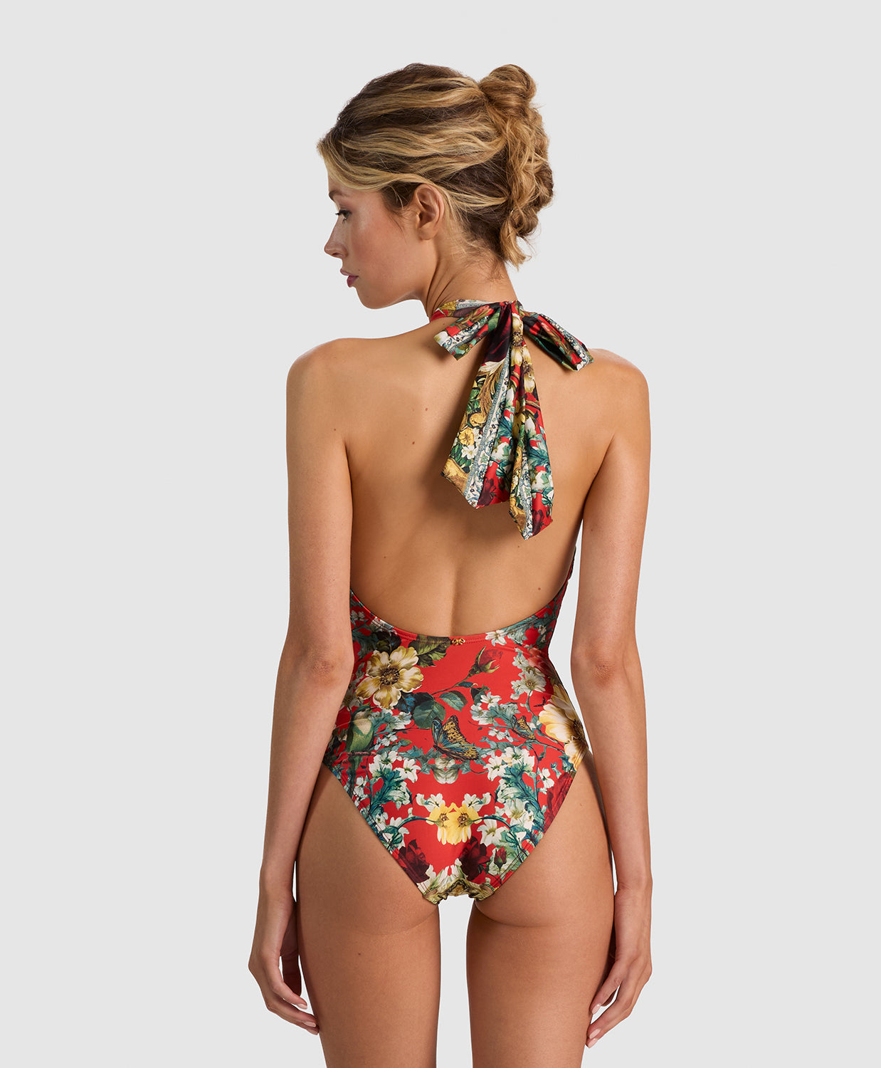 A woman shown from the back wearing a one-piece swimsuit with a red base and a colorful floral print, featuring a tie-neck design. The background is a solid light gray studio backdrop.