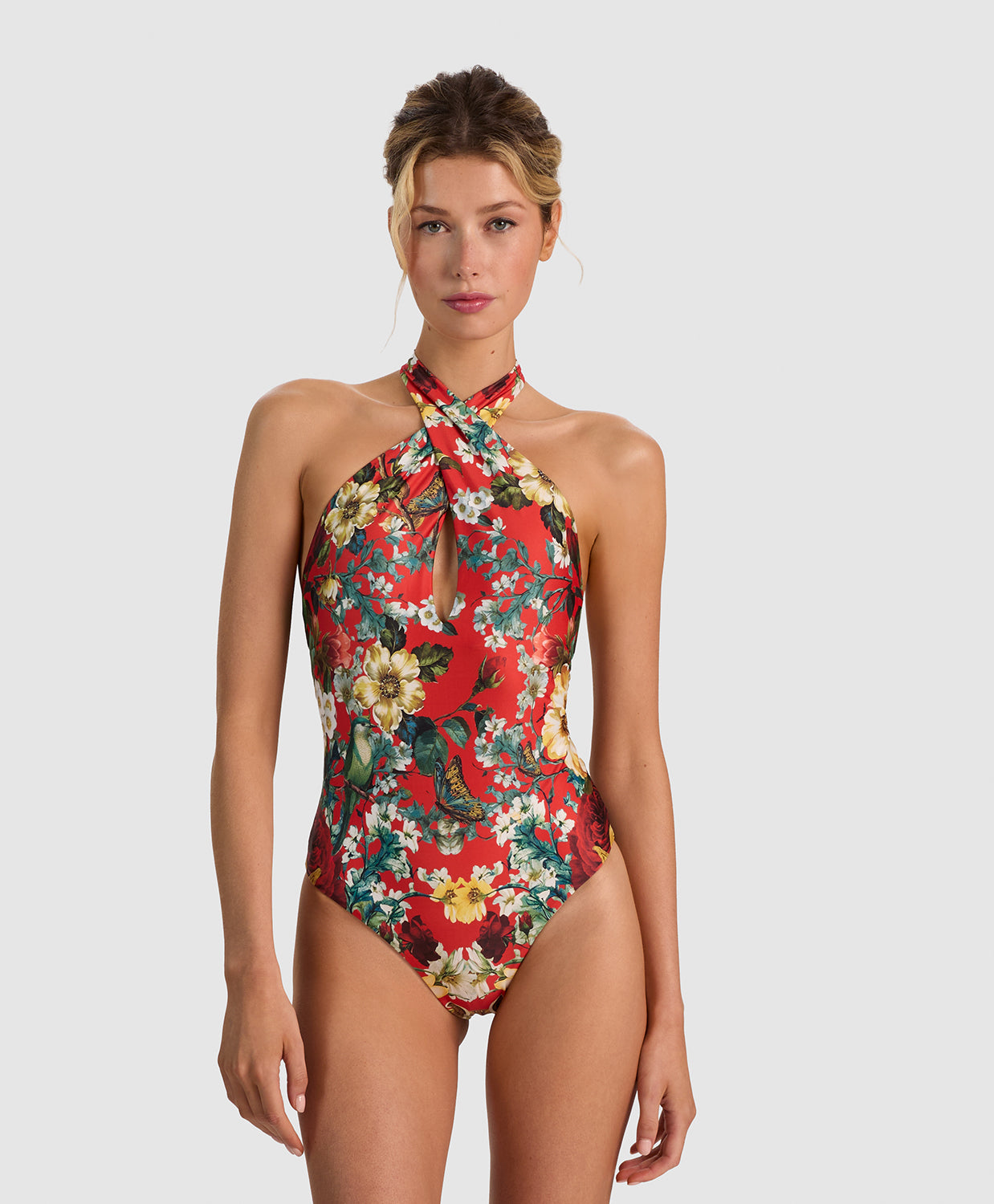 A woman modeling a red one-piece swimsuit with a vibrant floral print in yellow, white, and green, set against a solid light gray background.