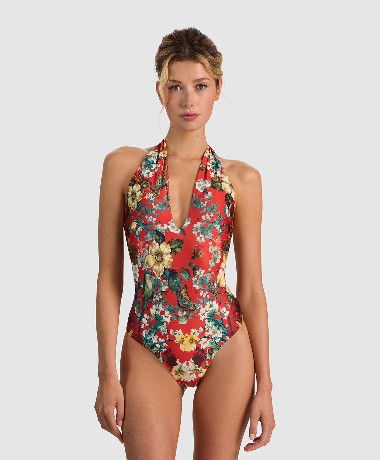 A woman modeling a red one-piece halter swimsuit with a colorful floral and bird print, set against a solid light gray background.