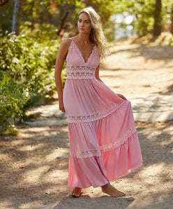 A blonde woman wearing a pink floor length dress standing on a dirt road near trees. 