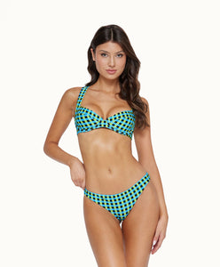 A brunette woman wearing a blue and green checkered bikini stands in front of a white wall. 