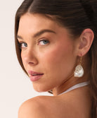 A woman wearing a white triangle top bikini and shell earrings standing against a white wall.