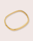 A gold square bracelet against a white wall.