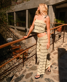 A woman wearing a beige and cream wavy knit two-piece set consisting of a cropped tank top and matching wide-leg pants, standing on a stone patio with a wooden railing.
