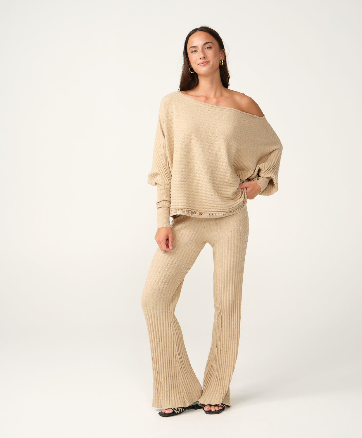 A woman modeling a matching beige knit two-piece set consisting of an off-the-shoulder long-sleeve top and ribbed wide-leg pants, photographed against a clean white studio background.
