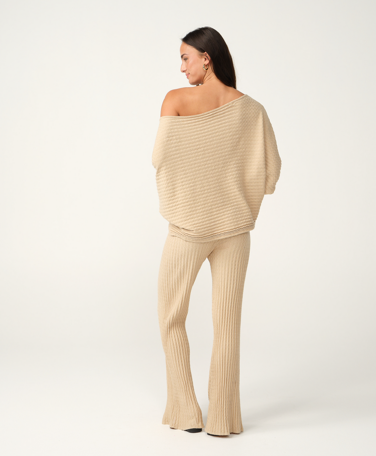 A woman modeling a matching beige knit two-piece set, featuring an off-the-shoulder cropped sweater and ribbed flare pants, shown from a rear three-quarter angle against a plain white studio background.