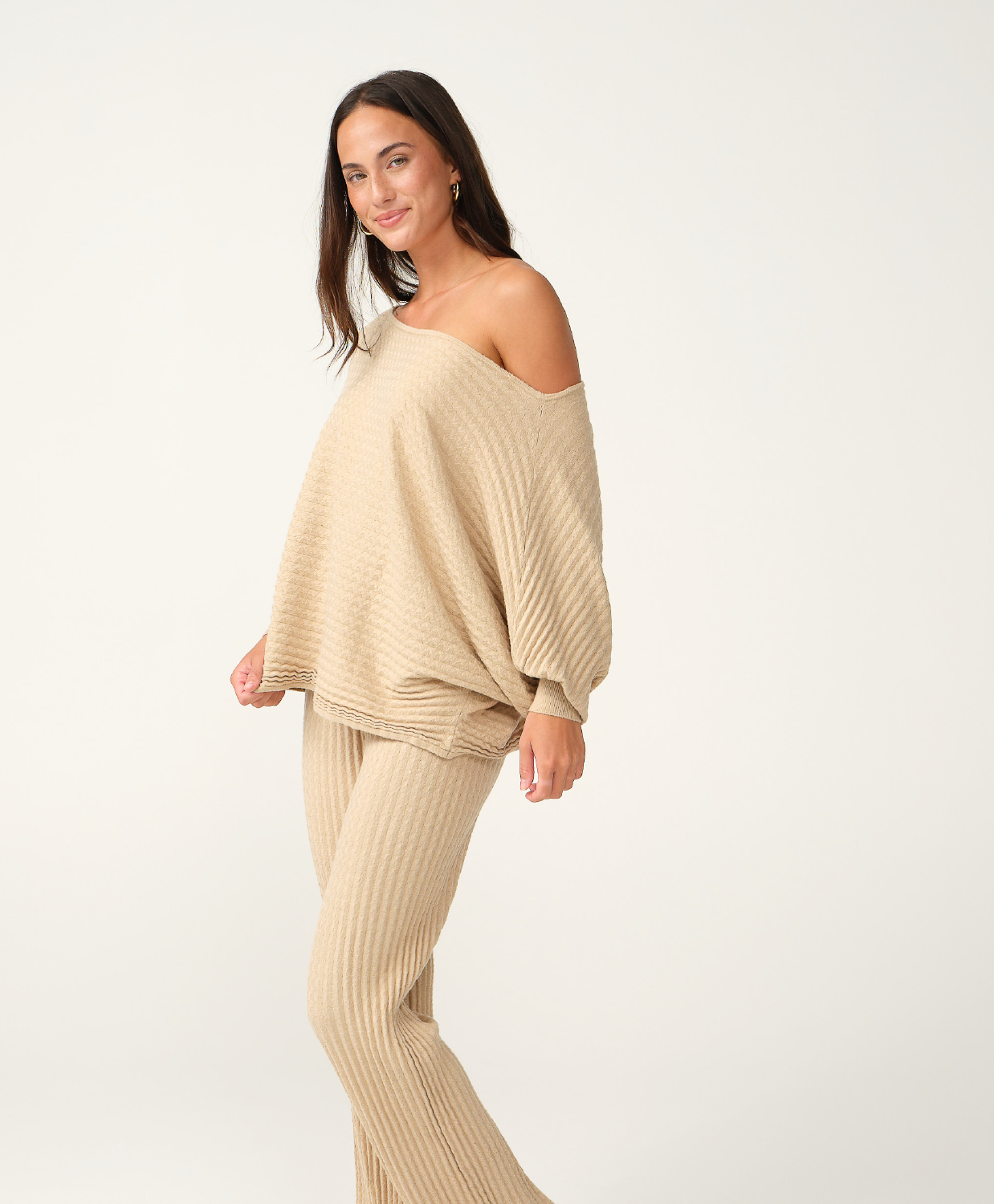 A woman modeling a matching beige ribbed knit lounge set consisting of a wide, off-the-shoulder sweater and ribbed pants, photographed against a clean white studio background.