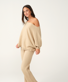 A woman modeling a matching beige ribbed knit lounge set consisting of a wide, off-the-shoulder sweater and ribbed pants, photographed against a clean white studio background.