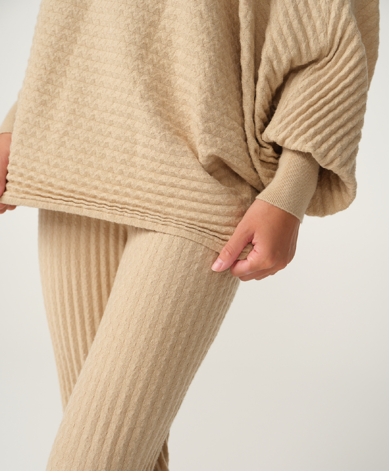 A close-up of a person wearing a beige ribbed knit sweater and matching ribbed knit pants against a plain light background.