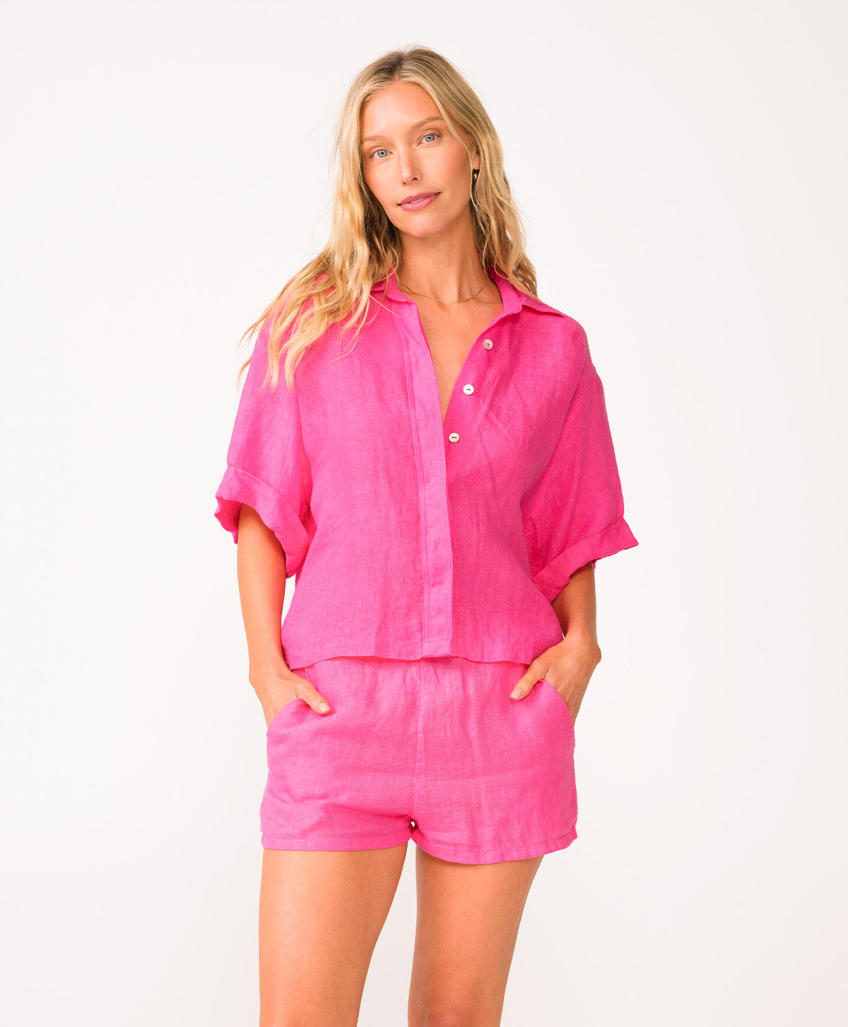 A woman wearing a bright pink linen shirt and matching shorts standing in front of a white wall.