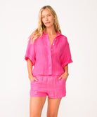 A woman wearing a bright pink linen shirt and matching shorts standing in front of a white wall.