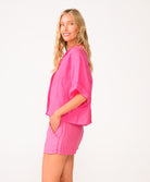 A woman wearing a bright pink linen shirt and matching shorts standing in front of a white wall.