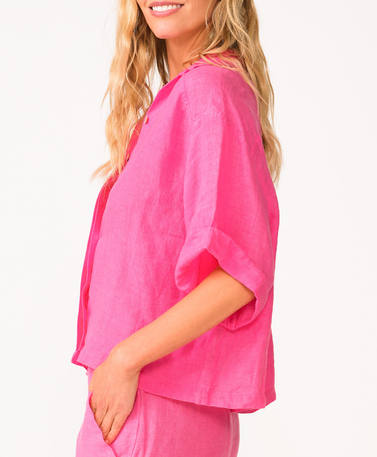 A woman wearing a bright pink linen shirt and matching shorts standing in front of a white wall.