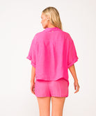 A woman wearing a bright pink linen shirt and matching shorts standing in front of a white wall.